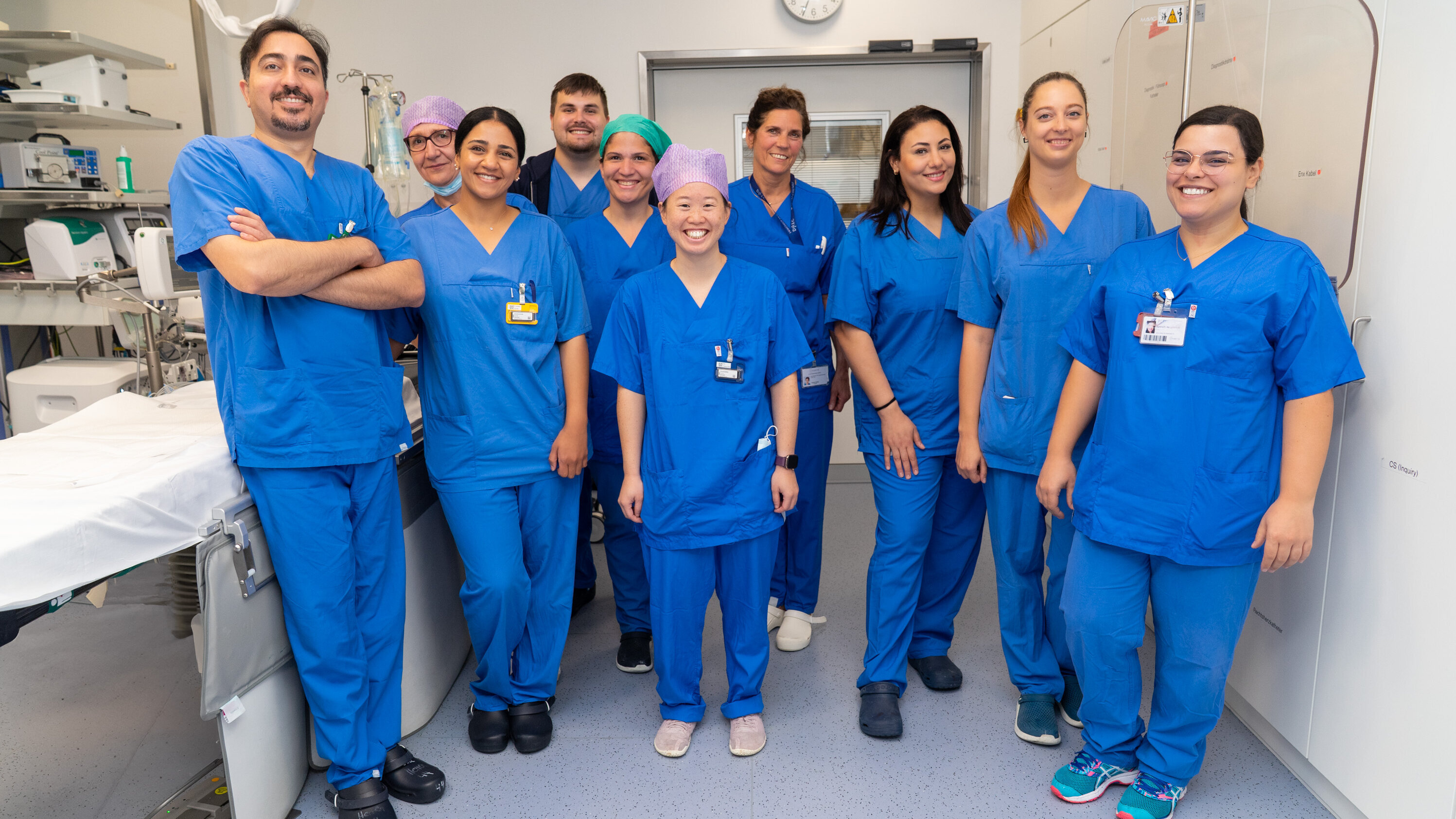 Eine Gruppe von zehn Personen in blauen Arzt- oder Pflegeuniformen steht lächelnd zusammen in einem modernen Behandlungsraum. Im Hintergrund sind medizinische Geräte und eine Uhr sichtbar. Alle Personen wirken freundlich und professionell.