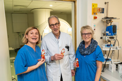 Drei lächelnde Personen in blauen Ärztekitteln stehen vor einem medizinischen Gerät. Sie halten jeweils einen Schokoladen-Weihnachtsmann in der Hand. Die Szene vermittelt eine freundliche und festliche Atmosphäre in einem Krankenhausumfeld.