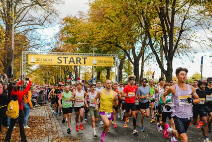 Eine Gruppe von Läufern startet beim Dresden Marathon. Unter einem Banner mit dem Wort "START" laufen die Teilnehmer in verschiedenen Sportoutfits auf einer von Bäumen gesäumten Straße. Die Atmosphäre ist lebhaft, mit Zuschauern, die die Läufer anfeuern.