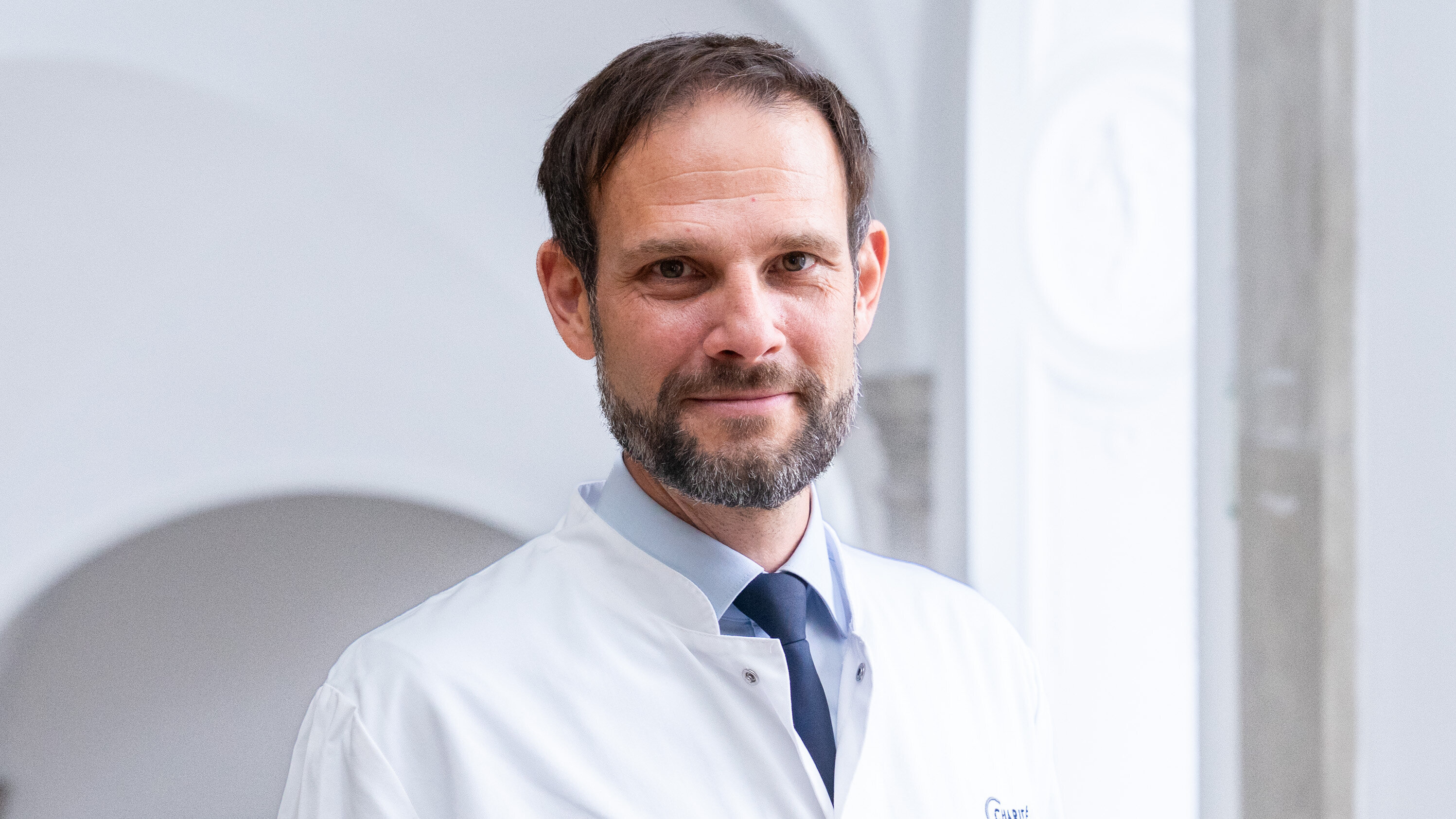 Ein Mann mit Bart trägt einen weißen Arztkittel und ein blaues Hemd. Er lächelt und steht vor einem hellen, neutralen Hintergrund, der eine angenehme Atmosphäre ausstrahlt.