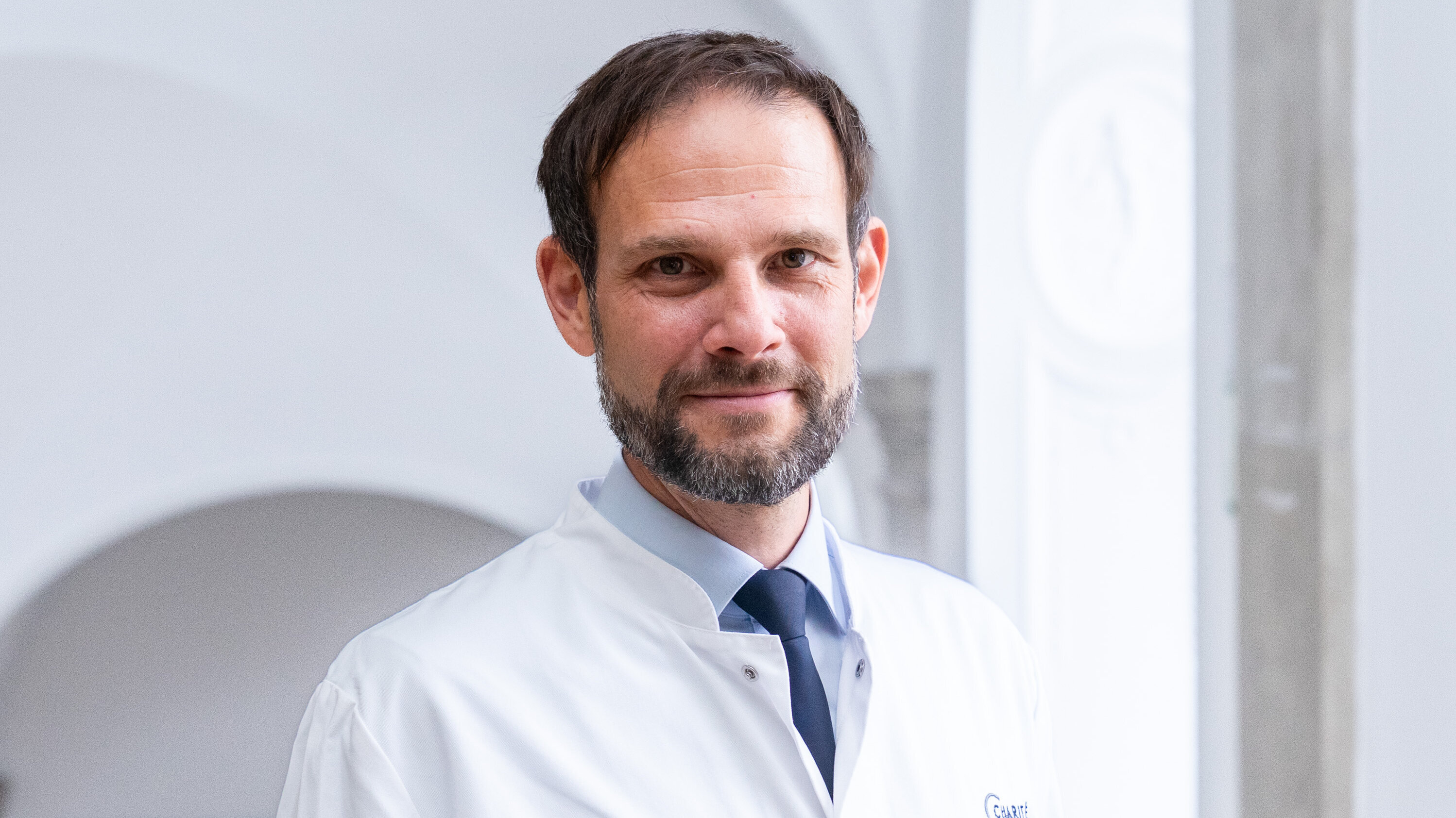 Ein Mann mit Bart trägt einen weißen Arztkittel und ein blaues Hemd. Er lächelt und steht vor einem hellen, neutralen Hintergrund, der eine angenehme Atmosphäre ausstrahlt.