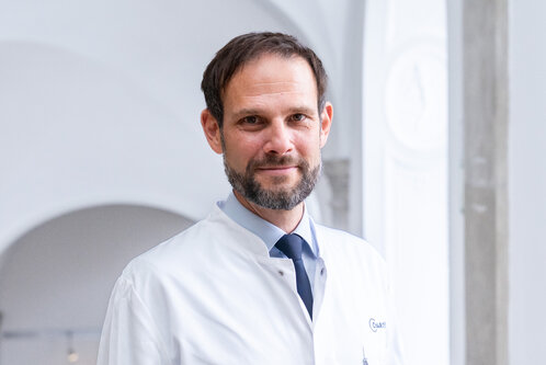 Ein Mann mit Bart trägt einen weißen Arztkittel und ein blaues Hemd. Er lächelt und steht vor einem hellen, neutralen Hintergrund, der eine angenehme Atmosphäre ausstrahlt.