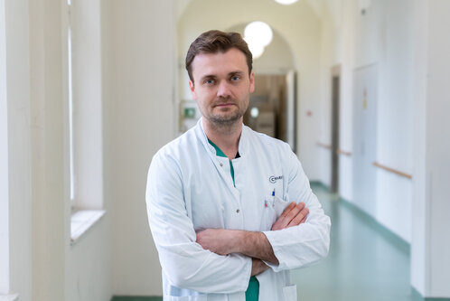 Ein Arzt in einem weißen Kittel steht mit verschränkten Armen in einem hellen Flur eines Krankenhauses. Sein Gesichtsausdruck vermittelt Professionalität und Entschlossenheit. Der Hintergrund zeigt helle Wände und Fenster, die Licht in den Raum lassen.