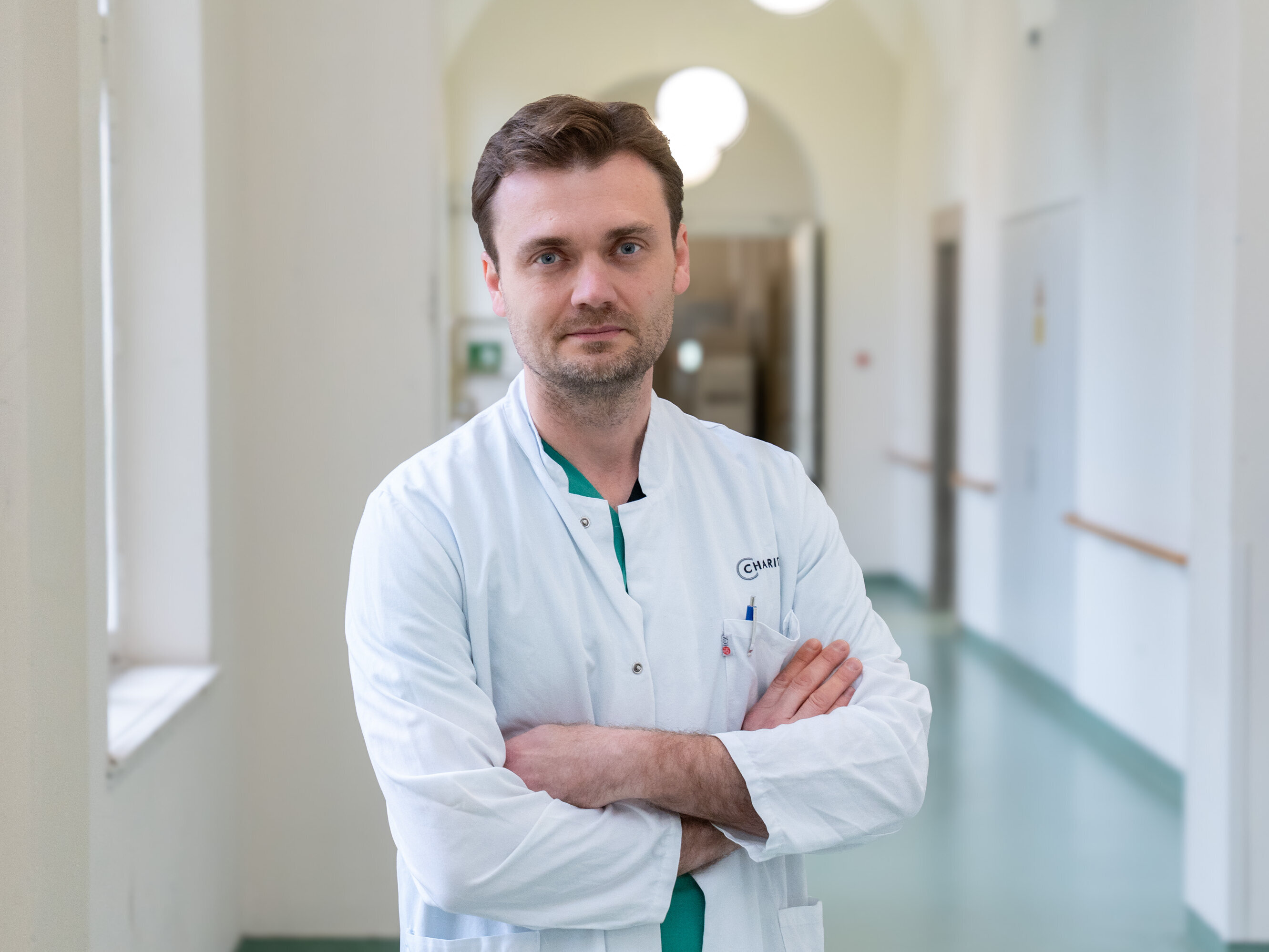 Ein Arzt in einem weißen Kittel steht mit verschränkten Armen in einem hellen Flur eines Krankenhauses. Sein Gesichtsausdruck vermittelt Professionalität und Entschlossenheit. Der Hintergrund zeigt helle Wände und Fenster, die Licht in den Raum lassen.
