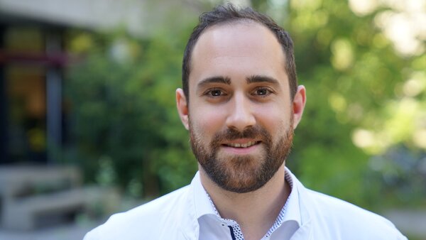 Ein junger Mann mit einem Bart und einem Lächeln trägt einen weißen Kittel. Er steht im Freien, vor einer unscharfen grünen Kulisse. Sein Gesicht ist deutlich sichtbar und vermittelt ein freundliches und professionelles Erscheinungsbild.