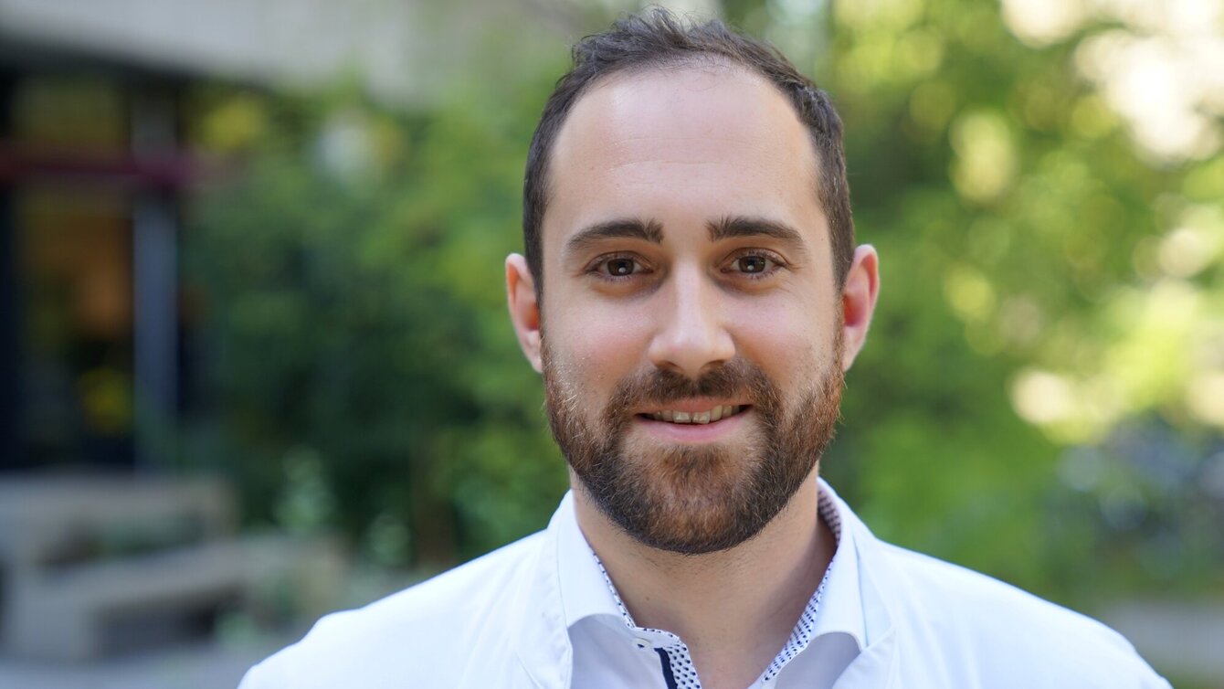 Ein junger Mann mit einem Bart und einem Lächeln trägt einen weißen Kittel. Er steht im Freien, vor einer unscharfen grünen Kulisse. Sein Gesicht ist deutlich sichtbar und vermittelt ein freundliches und professionelles Erscheinungsbild.