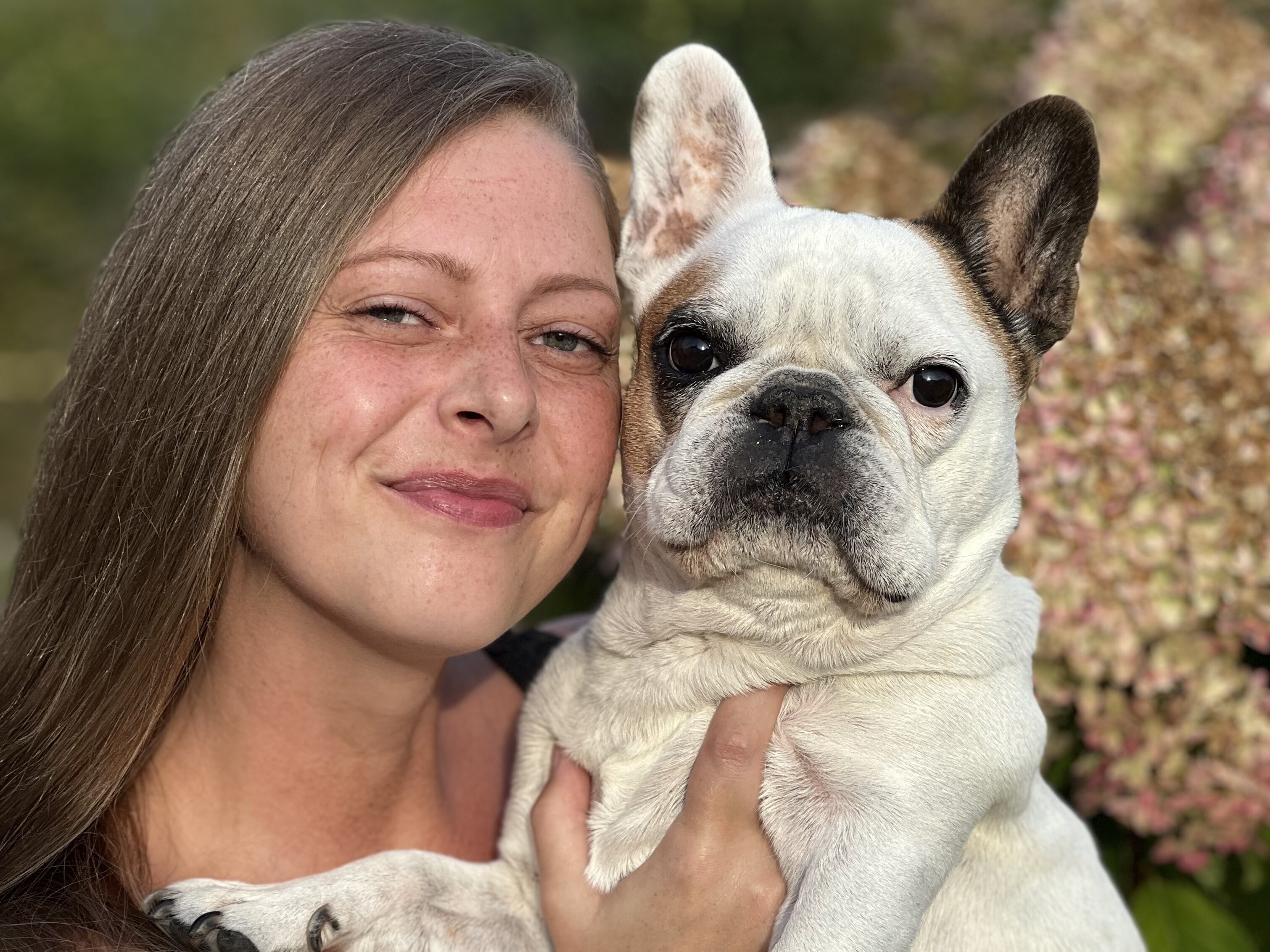 Eine Frau mit langen Haaren lächelt und hält einen französischen Bulldogg im Arm. Beide wirken glücklich und entspannt, umgeben von blühenden Pflanzen im Hintergrund.