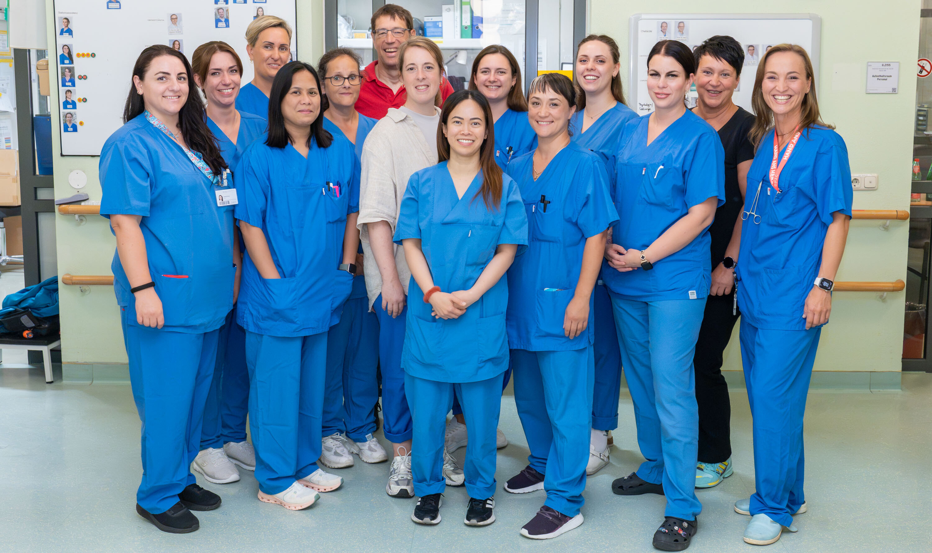 Eine Gruppe von 15 Personen in blauen Krankenhausuniformen steht zusammen in einem Innenraum. Sie lächeln und scheinen freundlich zu sein. Im Hintergrund sind Wände mit Informationsanzeigen zu sehen. Die Gruppe besteht aus verschiedenen Geschlechtern und Ethnien.