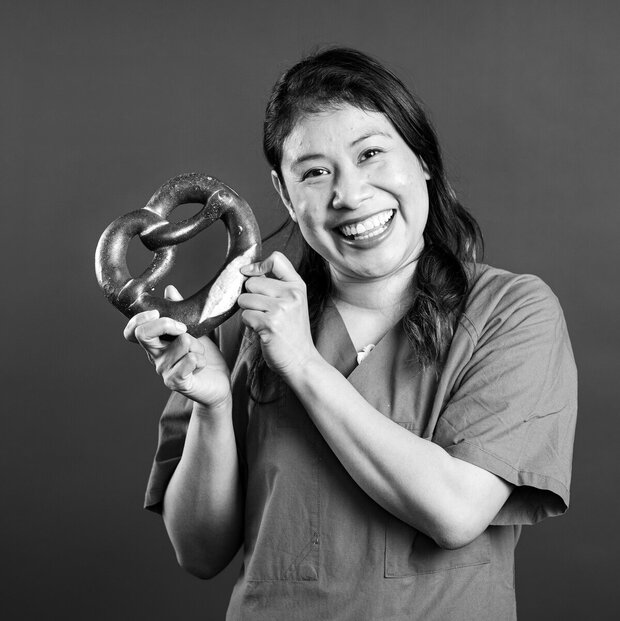 Eine lächelnde Frau hält eine weiche Brezel in der Hand und zeigt sie stolz. Sie trägt ein einfaches Oberteil und hat lange, dunkle Haare. Der Hintergrund ist einfarbig und macht die Brezel und ihr strahlendes Lächeln deutlich sichtbar.