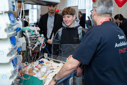 Ein junger Mann steht lächelnd vor einem Krankenbett, während ein älterer Mann in einem Fachshirt mit ihm spricht. Das Bett ist mit medizinischen Geräten und einem Puppenmodell für die Ausbildung ausgestattet. Im Hintergrund sind weitere Personen und farbige Ballons sichtbar.