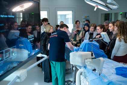 Eine Gruppe von Menschen beobachtet einen Arzt, der in einem Operationssaal eine medizinische Prozedur an einem Patienten durchführt. Der Raum ist modern ausgestattet, und eine Präsentation wird auf einem Monitor angezeigt. Die Anwesenden sind aufmerksam und nehmen Notizen.