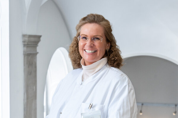 Eine lächelnde Frau in einem weißen Kittel steht in einem hellen Raum mit Bögen. Sie trägt eine Brille und hat lockige Haare. Ihre Haltung strahlt Freundlichkeit und Professionalität aus.