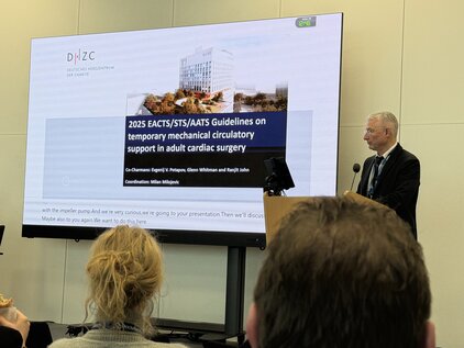 Ein Referent steht vor einer Präsentation, die die "2025 EACTS/STS/AATS Richtlinien zur temporären mechanischen Kreislaufunterstützung bei Erwachsenen mit Herzchirurgie" behandelt. Im Hintergrund sind Zuschauer zu sehen, die auf die Folien achten.