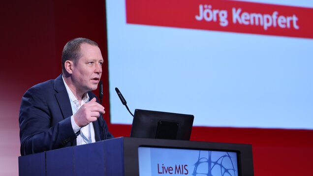 Ein Mann spricht auf einer Bühne bei einer Veranstaltung in Berlin. Er steht am Rednerpult und gestikuliert während seiner Präsentation. Im Hintergrund ist ein Banner mit dem Namen „Jörg Kempfert“ zu sehen. Der Kontext vermittelt eine professionelle Diskussion oder Konferenz.