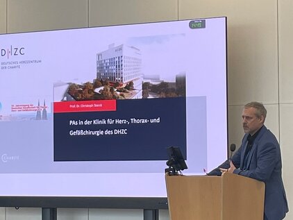Ein Vortragender steht neben einem Bildschirm, auf dem Informationen über das Deutsche Zentrum der Herz-Kreislaufmedizin (DHZC) zu sehen sind. Der Fokus liegt auf der Präsentation zu Patienten in der Klinik für Herz-, Thorax- und Gefäßchirurgie.