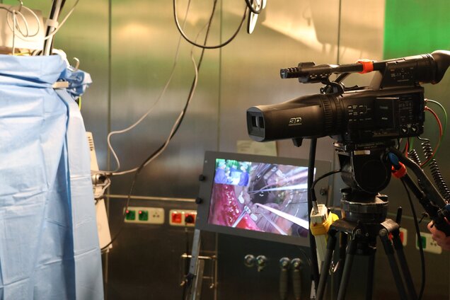 Eine Videokamera steht in einem Operationsteam mit einem Bildschirm, der eine nahaufgenommene Ansicht von chirurgischen Instrumenten zeigt. Der Raum wirkt steril und professionell, mit medizinischem Equipment im Hintergrund.