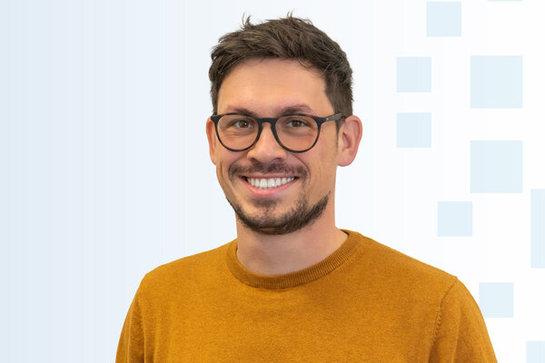 Ein lächelnder Mann mit Brille trägt einen orangefarbenen Pullover. Im Hintergrund sind sanfte geometrische Muster in Hellblau zu sehen. Der Ausdruck des Mannes vermittelt Freundlichkeit und Offenheit.