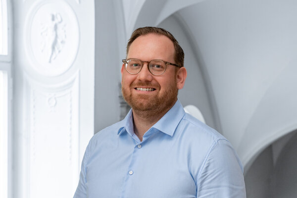 Ein Mann mit Brille lächelt freundlich in die Kamera. Er trägt ein hellblaues Hemd und steht vor einem hellen Hintergrund mit architektonischen Details.