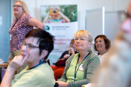 Die Aufnahme zeigt eine Gruppe von Menschen, die an einer Veranstaltung teilnehmen. Einige Teilnehmer hören aufmerksam zu, während andere miteinander sprechen. Eine Frau in der Vordergrund trägt eine Brille und hat blonde Haare. Im Hintergrund ist ein Plakat mit einer kinderfreundlichen Botschaft zu sehen.