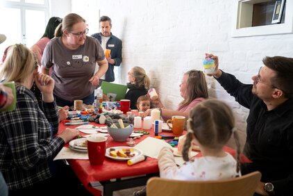 Eine Gruppe von Menschen sitzt an einem Tisch, der mit Bastelmaterialien, Getränken und Snacks bedeckt ist. Eine Frau spricht aktiv mit den Anwesenden, während Kinder und Erwachsene gemeinsam kreativ tätig sind und verschiedene Gegenstände gestalten.