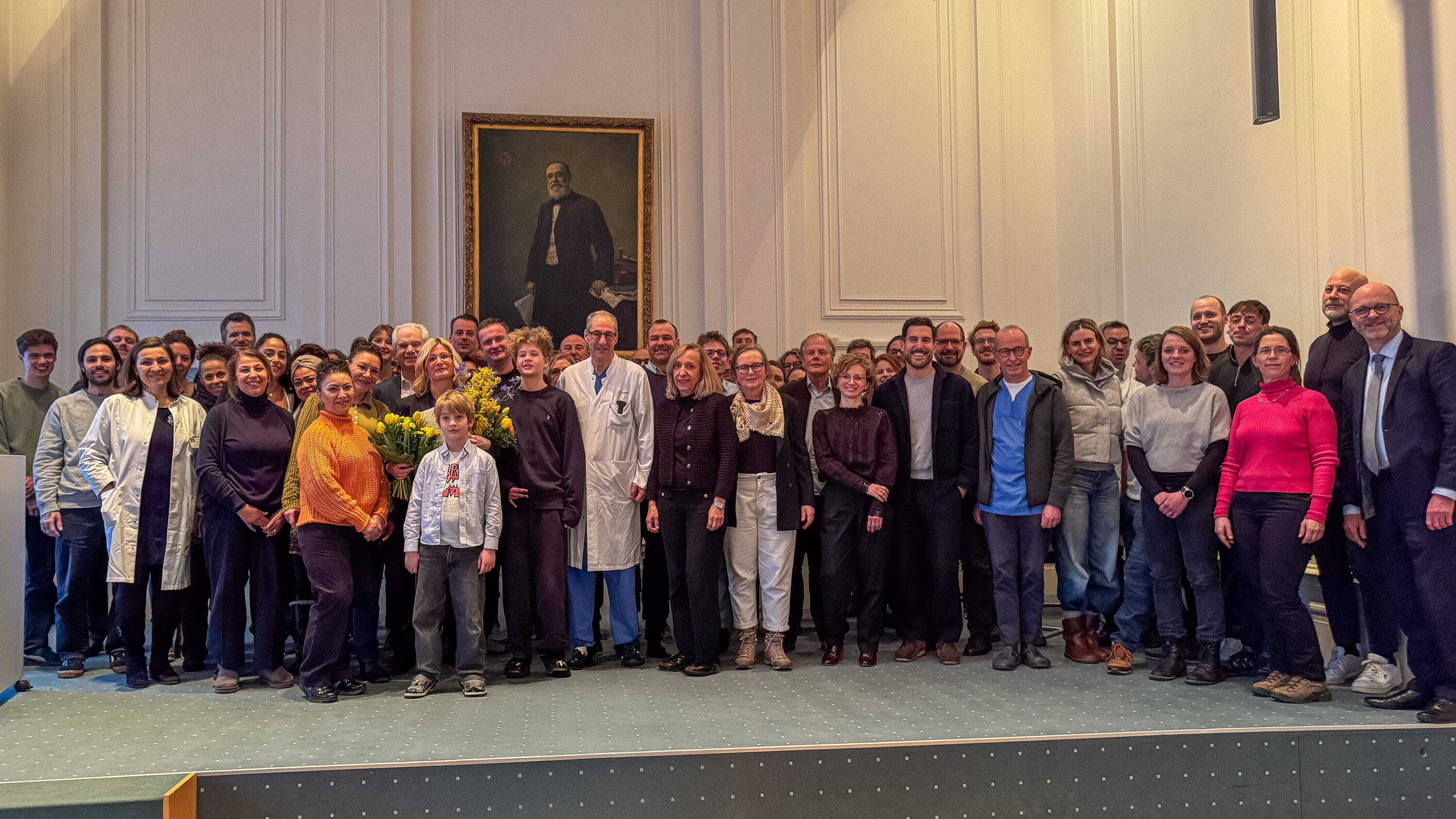 Eine große Gruppe von etwa 50 Personen steht in einem Raum mit einer hellen Wand. Einige Personen tragen weiße Laborkittel oder medizinische Kleidung. Eine Frau hält einen Strauß mit gelben Blumen in der Mitte der Gruppe. Im Hintergrund hängt ein Porträt an der Wand.