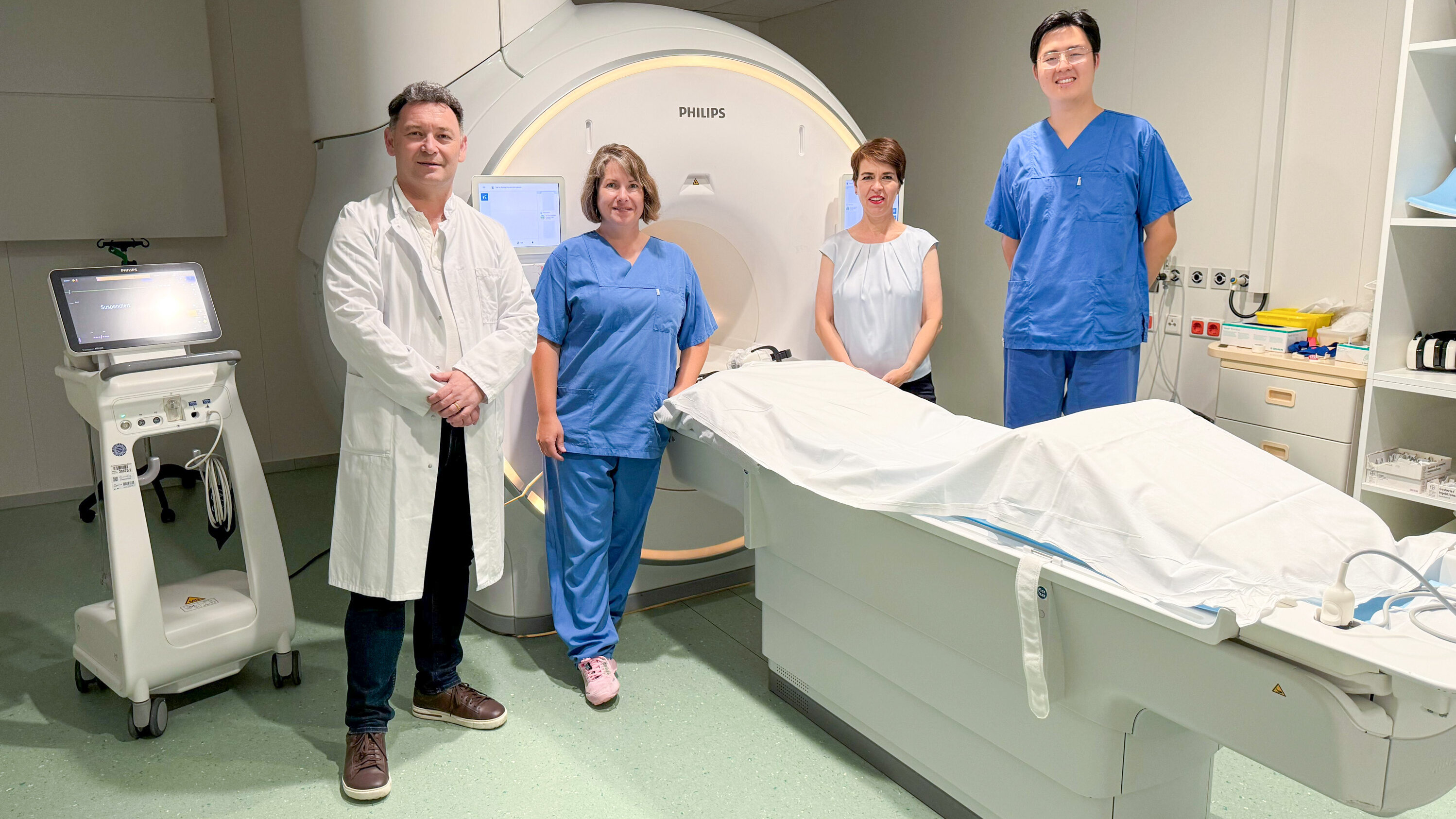 Vier medizinische Fachkräfte stehen in einem Untersuchungsraum. Hinter ihnen befindet sich ein modernes MRI-Gerät. Die Personen tragen Arbeitskleidung: ein Arzt in einem weißen Kittel, zwei Mitarbeiter in blauen Scrubs und eine weitere Person in einem hellen Oberteil. Ein Untersuchungstisch ist ebenfalls sichtbar.