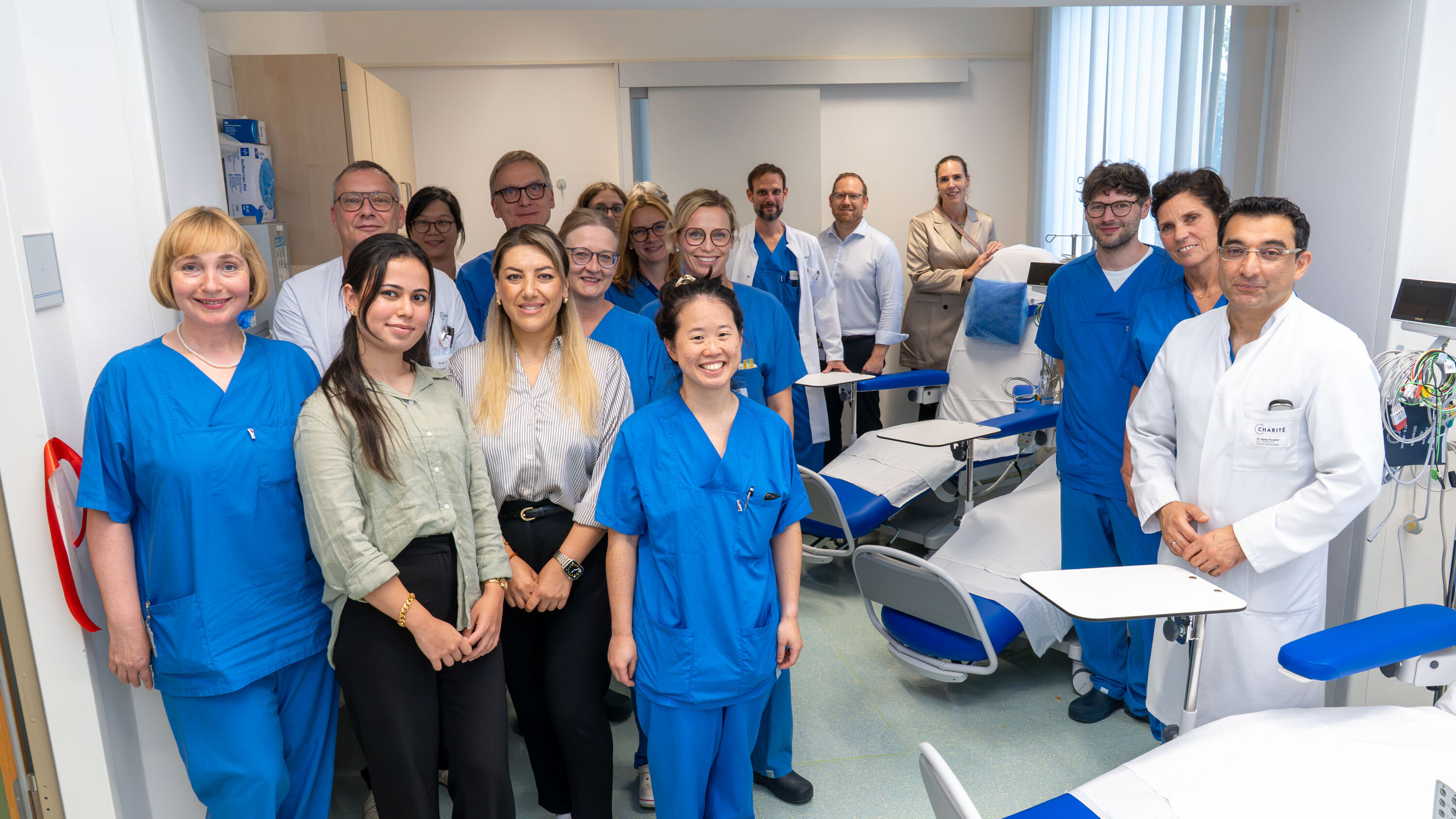 Eine Gruppe von 15 Personen steht in einer Klinik, gekleidet in blaue Arbeitskleidung. Im Hintergrund sind medizinische Geräte und Untersuchungsstühle zu sehen. Die Atmosphäre wirkt freundlich und kollegial. Einige Personen lächeln in die Kamera.