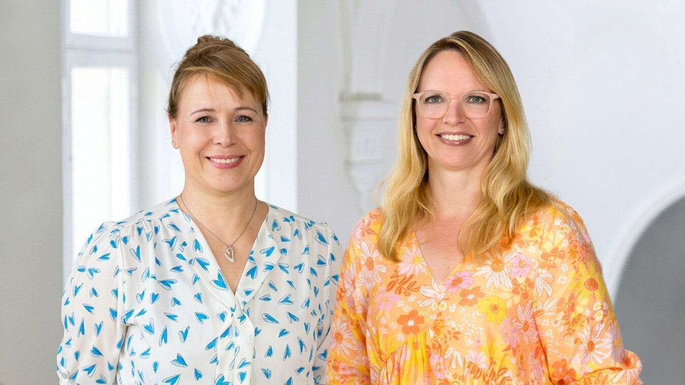 Zwei Frauen stehen in einem hellen Raum miteinander. Die erste trägt ein bläuliches, gemustertes Hemd, die zweite ein leuchtend orange-blühendes Kleid. Beide lächeln freundlich in die Kamera und strahlen Selbstbewusstsein aus.