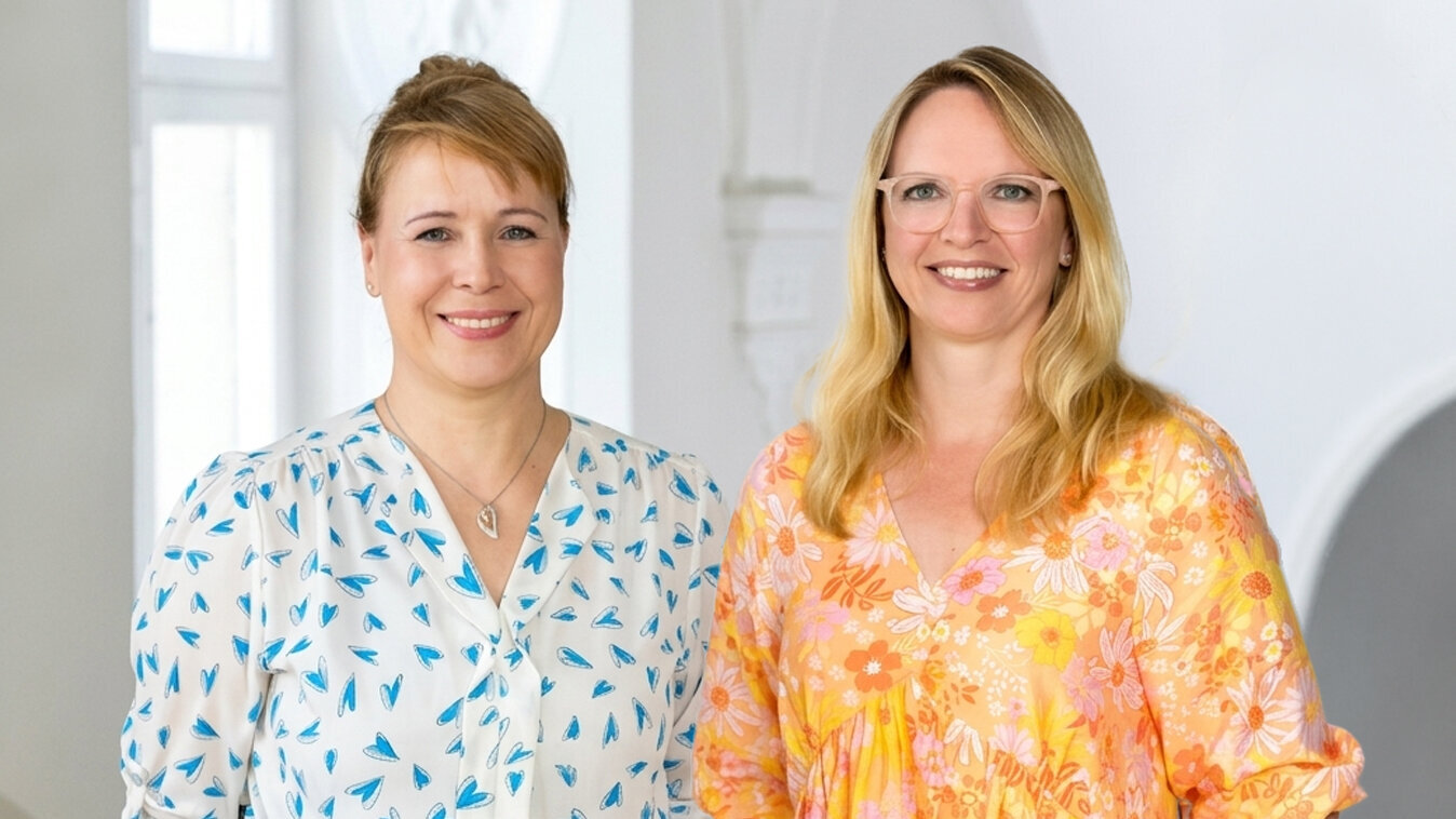Zwei Frauen stehen in einem hellen Raum miteinander. Die erste trägt ein bläuliches, gemustertes Hemd, die zweite ein leuchtend orange-blühendes Kleid. Beide lächeln freundlich in die Kamera und strahlen Selbstbewusstsein aus.