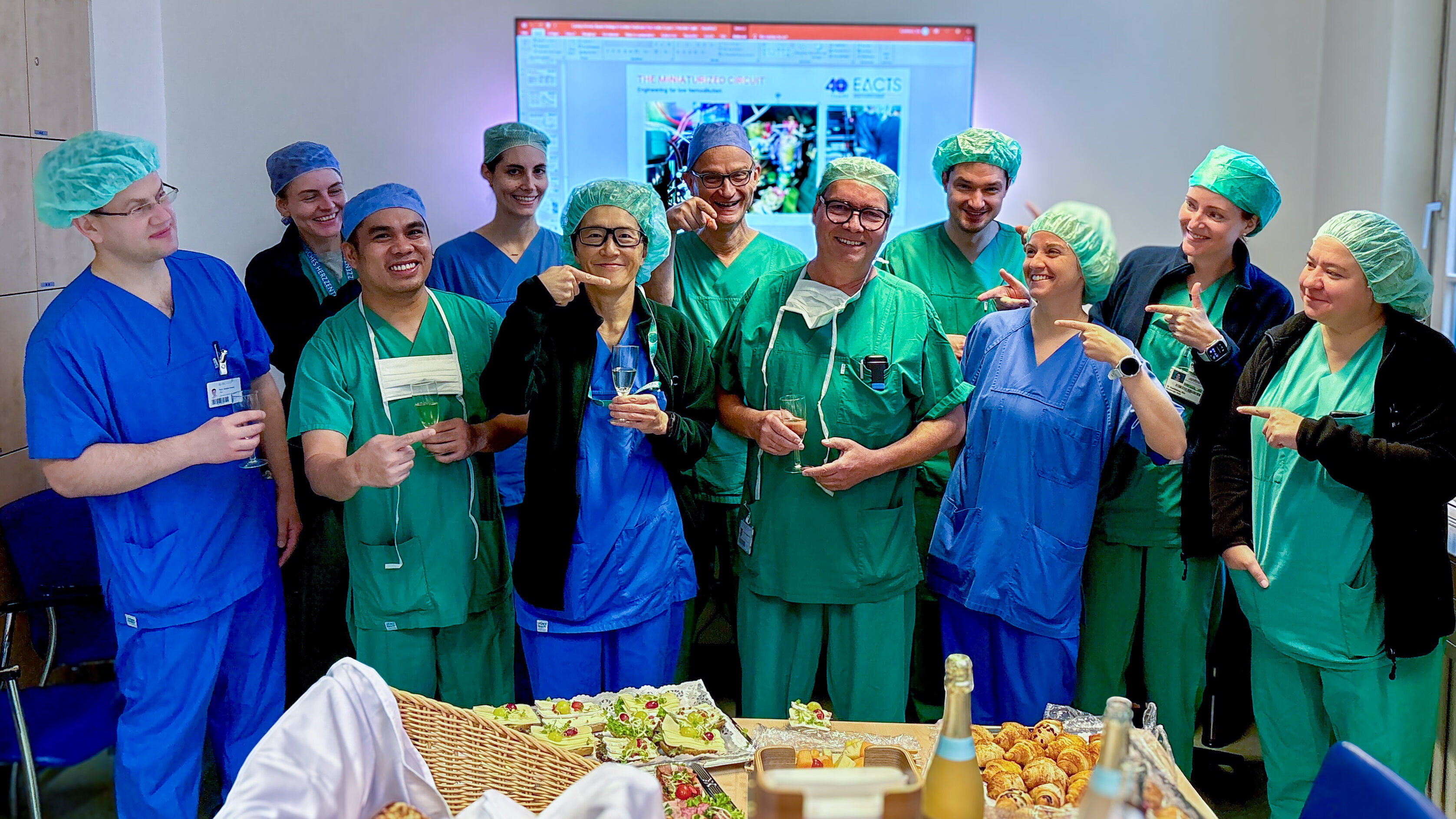 Eine Gruppe von elf Personen in OP-Kleidung und Haarnetzen steht zusammen und lächelt. Sie halten Getränke in den Händen und zeigen mit den Händen auf etwas im Hintergrund. Auf einem Tisch sind verschiedene Snacks und Getränke angerichtet.