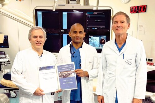 Drei Ärzte in weißen Kitteln stehen in einem modernen Krankenhausraum. Sie halten Zertifikate für den Preis für innovative Lehre 2025. Im Hintergrund sind medizinische Geräte und Bildschirme sichtbar. Alle drei lächeln stolz in die Kamera.