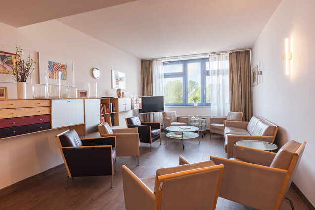 Ein moderner, hell gestalteter Raum mit Holzfußboden. In der Mitte stehen ein Glastisch und mehrere mehrfarbige Sessel. An der Wand hängen Bilder, und große Fenster lassen Tageslicht hinein. Ein Regal und ein TV-Gerät sind ebenfalls im Raum.