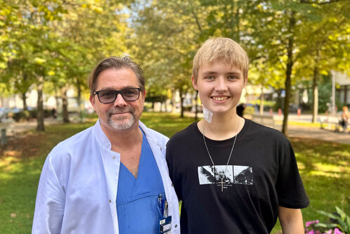 Ein männlicher Arzt mit Brille in medizinischer Kleidung steht neben einem jungen Mann, der ein schwarzes T-Shirt trägt. Beide lächeln und befinden sich in einem Park mit Bäumen und Blumen im Hintergrund.