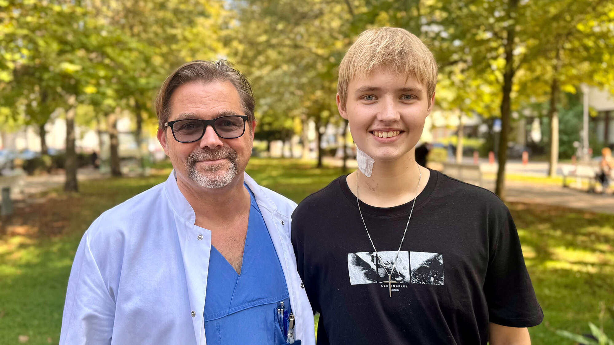Ein männlicher Arzt mit Brille in medizinischer Kleidung steht neben einem jungen Mann, der ein schwarzes T-Shirt trägt. Beide lächeln und befinden sich in einem Park mit Bäumen und Blumen im Hintergrund.