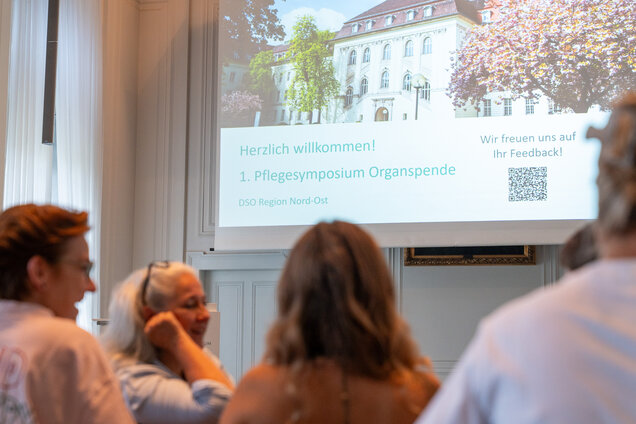 Ein Bildschirm zeigt die Willkommensnachricht für das 1. Pflegesymposium zur Organspende. Darunter steht, dass sich die DSO Region Nord-Ost über Feedback freut. Im Vordergrund sind Personen zu sehen, die miteinander sprechen.