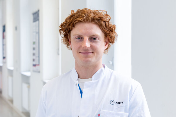 Ein junger Mann mit lockigem, rotem Haar trägt einen weißen Laborkittel mit dem Schriftzug "Charité". Er steht in einem hellen, modernen Flur und lächelt freundlich in die Kamera.