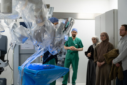 Ein medizinisches Team steht in einem Operationssaal, während ein Roboterarm, der in Plastikhülle verpackt ist, vorbereitet wird. Ein Arzt in grüner Kleidung schaut auf den Roboter, während drei weitere Personen aufmerksam zuschauen.