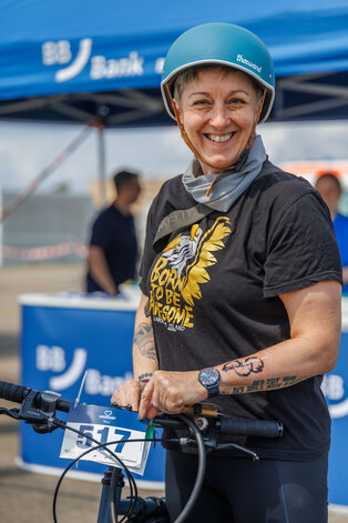 Eine lächelnde Frau trägt einen blauen Helm und ein schwarzes T-Shirt mit der Aufschrift "Born to be Awesome". Sie steht neben einem Fahrrad und hat eine Startnummer am Lenker befestigt. Im Hintergrund ist ein Veranstaltungszelt sichtbar.