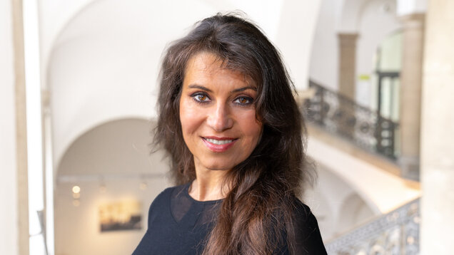 Eine Frau mit langen, dunklen Haaren lächelt in die Kamera. Sie trägt ein schwarzes Oberteil und steht in einer hellen Umgebung mit architektonischen Details im Hintergrund.