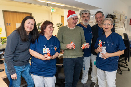 Sechs Personen stehen in einem Raum und lächeln. Sie tragen blaue Arbeitskleidung und halten bunte Schokoladenfiguren in Form von Weihnachtsmännern. Eine Person trägt einen Weihnachtsmann-Hut. Die Atmosphäre wirkt freundlich und festlich.