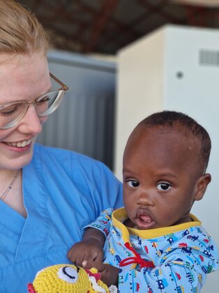 Eine junge Frau mit Brille lächelt freundlich, während sie ein Kleinkind in ihren Armen hält. Das Kind trägt einen bunten Schlafanzug und schaut direkt in die Kamera. Die Szene vermittelt eine warme, fürsorgliche Atmosphäre zwischen den beiden.