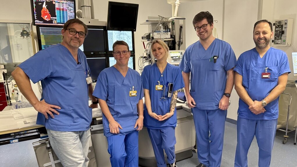 Fünf medizinische Fachkräfte in blauen Scrubs stehen in einem Krankenhauszimmer. Sie lächeln und posieren gemeinsam vor einem medizinischen Gerät. Die Szene vermittelt Teamarbeit und Professionalität im Gesundheitsbereich.