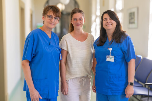 Drei Frauen stehen in einem hellen Flur. Zwei von ihnen tragen blaue Krankenhauskleidung, die dritte trägt ein helles, lockeres Oberteil und kurze Hosen. Alle drei lächeln freundlich. Im Hintergrund sind Stühle zu sehen.