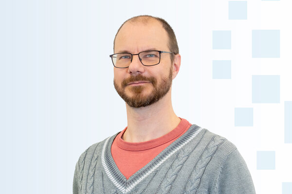 Ein Mann mit Brille und einem kurzen Bart lächelt leicht. Er trägt einen grauen Pullover mit einem roten T-Shirt darunter. Der Hintergrund ist hell mit geometrischen Formen.