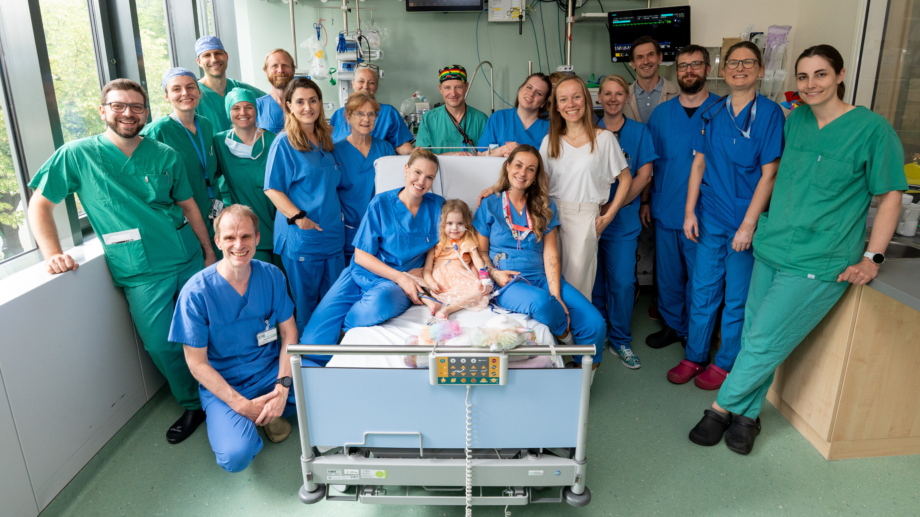 Eine Gruppe von über 20 medizinischen Fachkräften, darunter Ärzte und Pflegekräfte, posiert lächelnd um ein Krankenbett, in dem ein Kind mit einer Umarmung sitzt. Alle sind in grünen und blauen Krankenhauskleidern gekleidet. Die Szene vermittelt eine Atmosphäre der Unterstützung und Fürsorge.