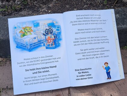 Aufgeschlagenes Kinderbuch mit Illustrationen und Text. Es zeigt ein Krankenhauszimmer mit einem Kind und einem Roboter. Der Text erzählt von einer Geschichte, in der das Kind und der Roboter interagieren. Am unteren Rand steht: "Eine Geschichte für Maloa, in voller Liebe von deiner Oma."
