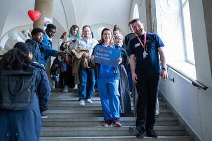 Eine Gruppe von Menschen steht auf einer Treppe. Eine Frau in blauer Arbeitskleidung hält ein Schild mit der Aufschrift „Führung Hybrid OP“ in die Kamera. Andere Personen begleiten sie und schauen in Richtung der Kamera.