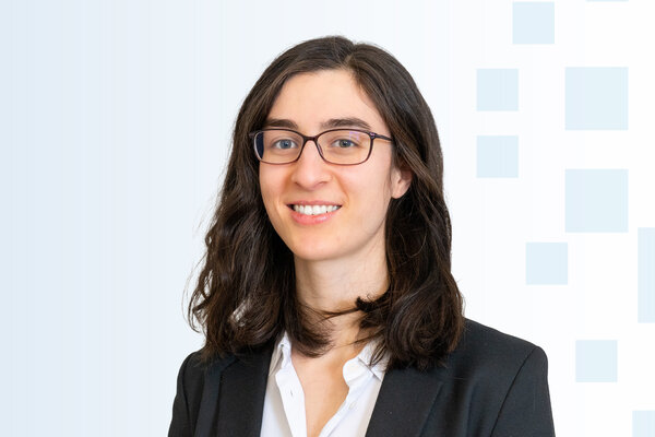 Eine Frau mit langen, dunklen Haaren und einer schwarzenjacke lächelt in die Kamera. Sie trägt eine weiße Bluse und Brillen. Der Hintergrund ist hell und grafisch strukturiert, mit sanften, abgestuften Farben.