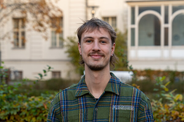 Ein junger Mann mit einem freundlichen Lächeln steht in einem Park. Er trägt ein kariertes Hemd in grünen und blauen Farbtönen. Im Hintergrund sind einige Bäume und ein Gebäude zu sehen, das die herbstliche Atmosphäre vermittelt.