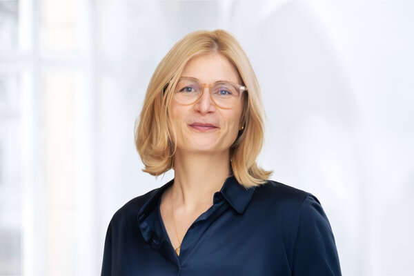 Eine Frau mit blonden, schulterlangen Haaren und einer dunklen Bluse lächelt in die Kamera. Sie trägt eine modische Brille. Der Hintergrund ist hell und unscharf, was den Fokus auf die Frau lenkt.